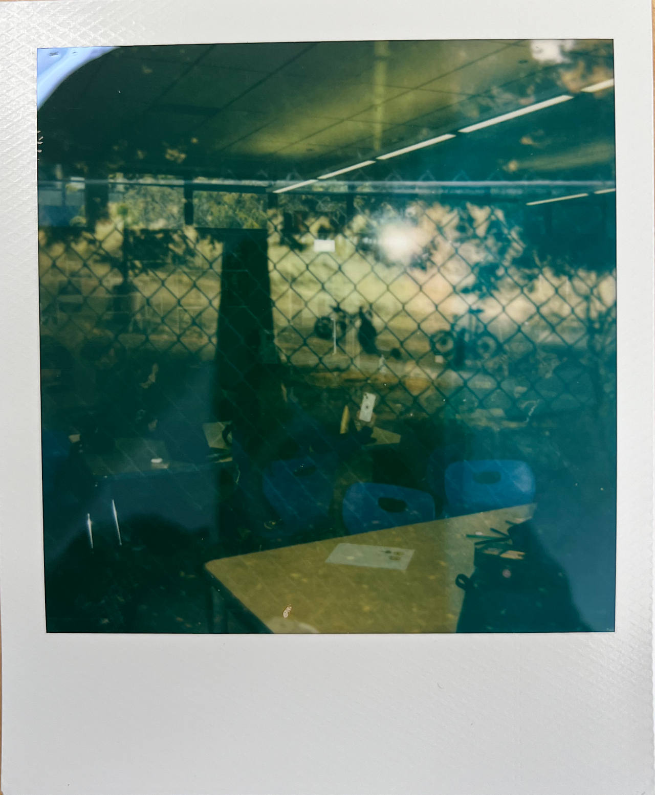 Dim industrial interior seen through chain-link fence, with bright lights and a table in foreground.