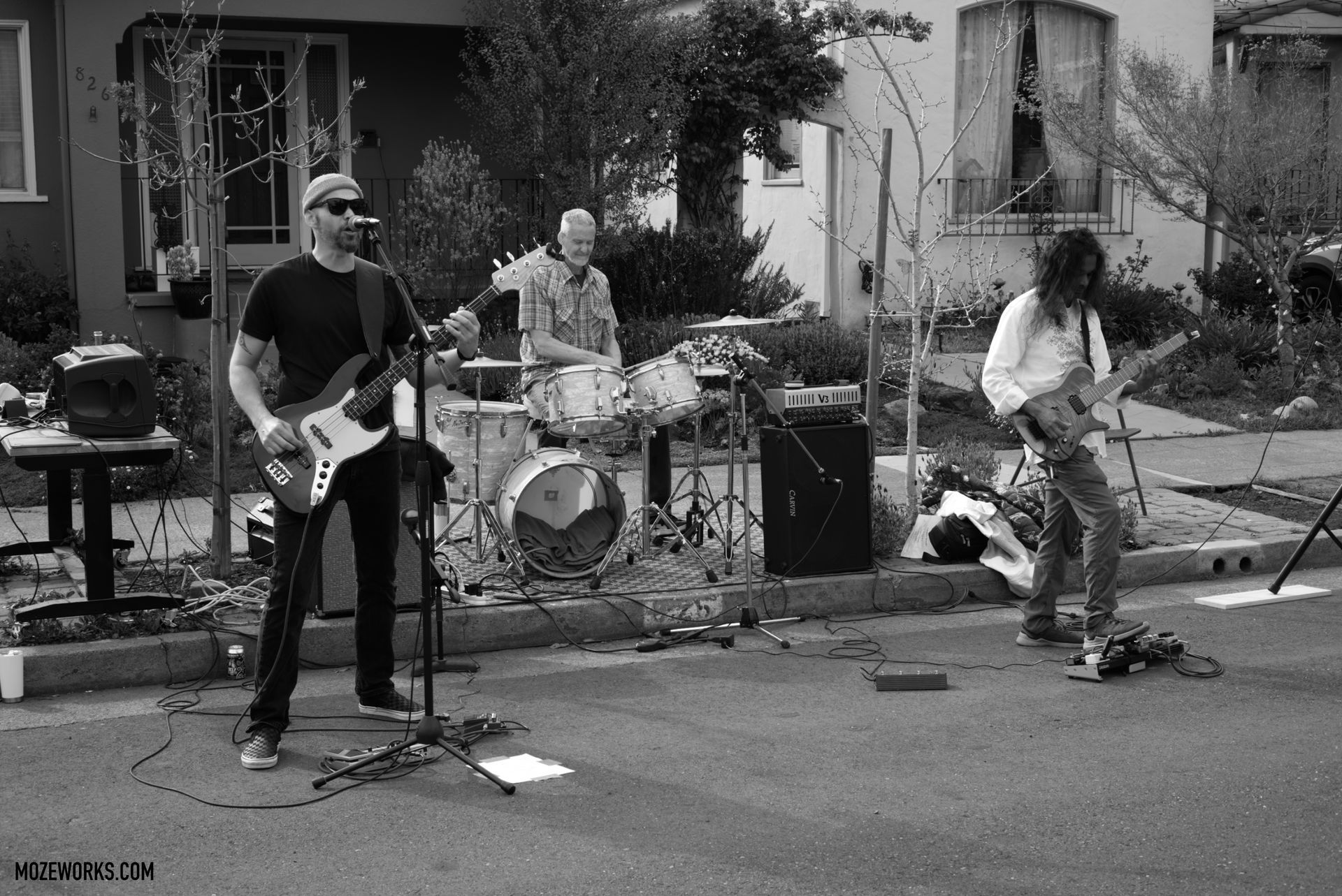 Three musicians performing outdoors in a front yard, with guitars, drums, and microphones.