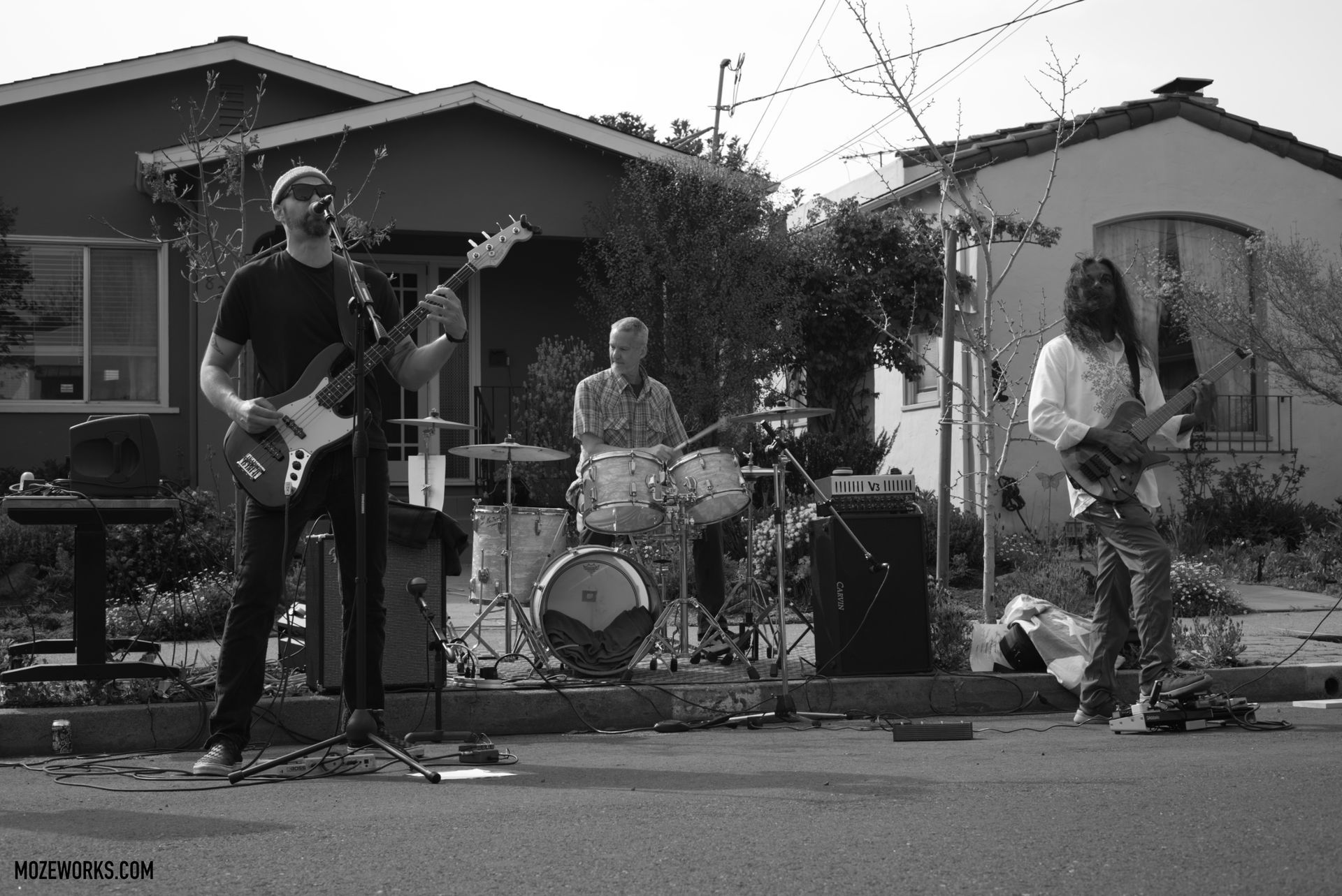 Outdoor band performing in a residential street, with guitarists and drummer in front of houses.