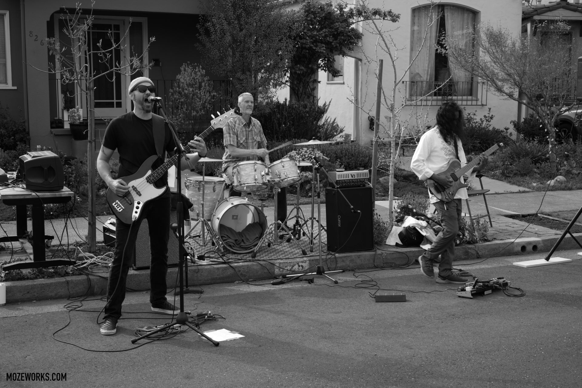 Band playing guitars and drums outdoors on a residential street, with houses and trees behind them.