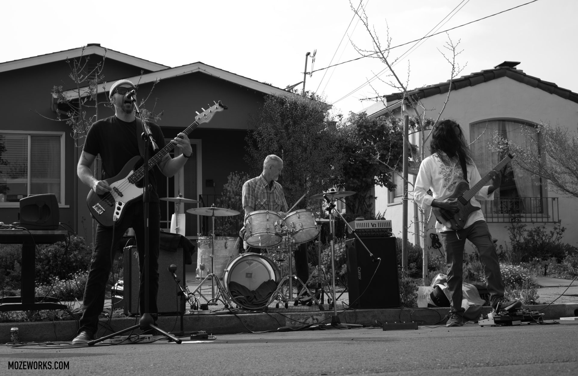 Three musicians playing instruments outdoors in front of houses, in black and white.