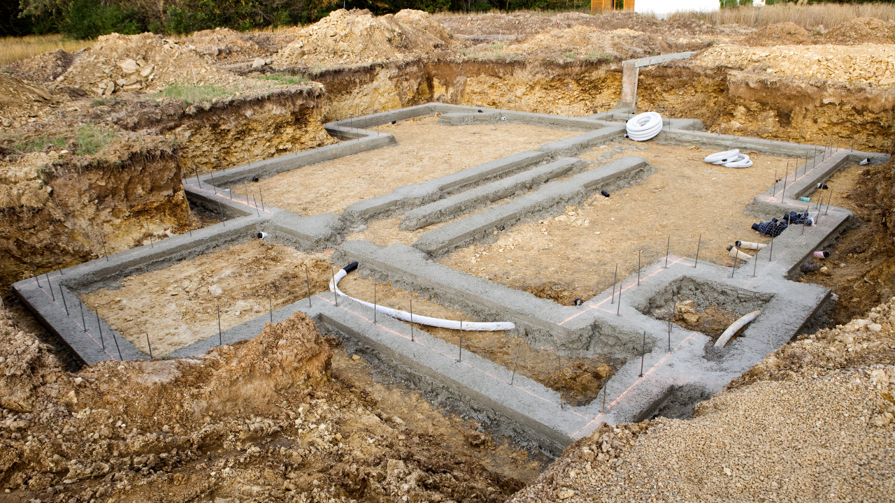Concrete foundation layout in a dirt excavation site, with gray footings and pipes forming a building plan