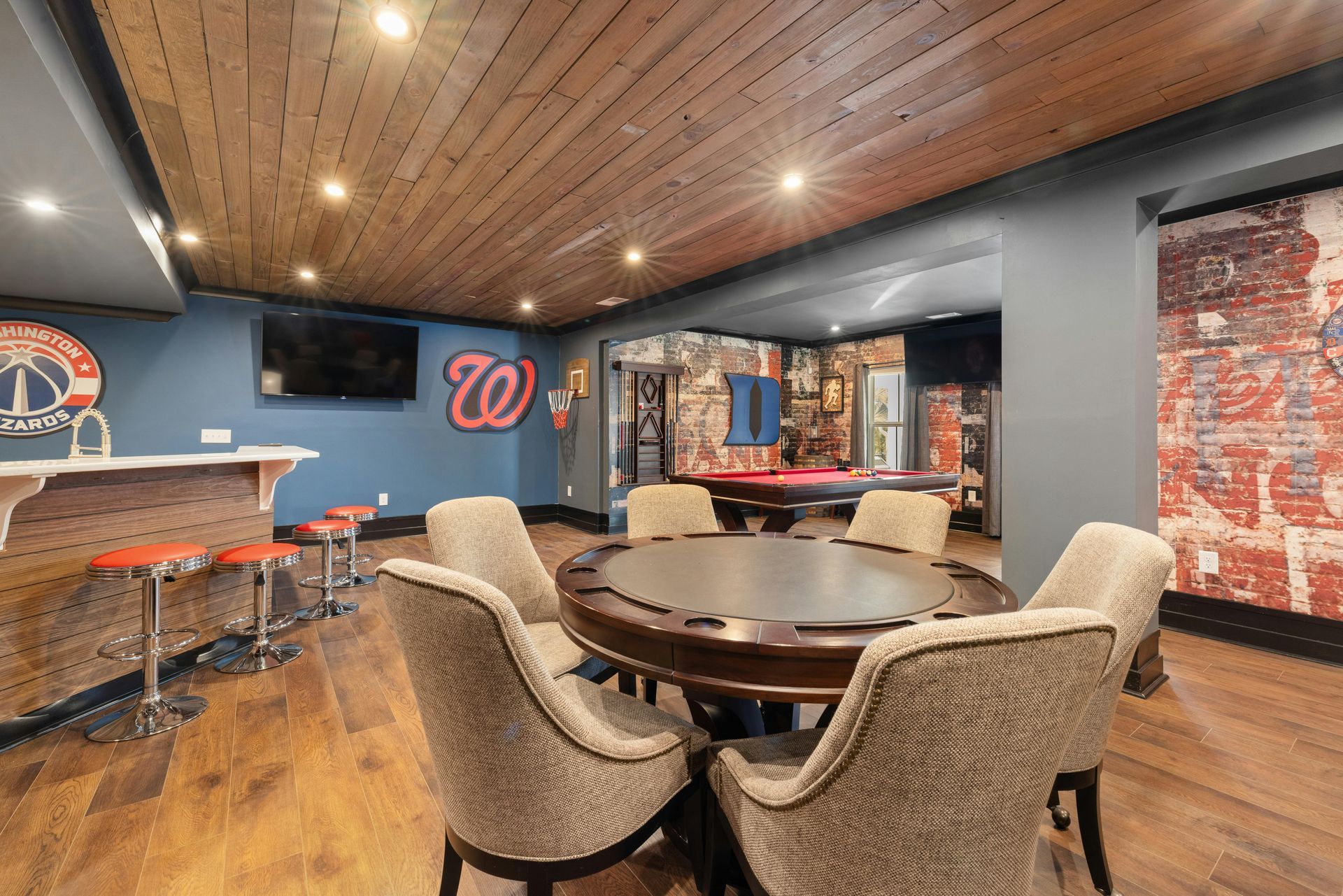 Modern lounge with round table, upholstered chairs, bar stools, TV, and wood-paneled ceiling