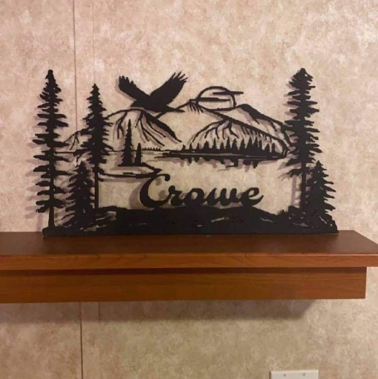 Black metal wall art of mountains, trees, and a bird, with “Coeur” on a wood shelf against a beige wall