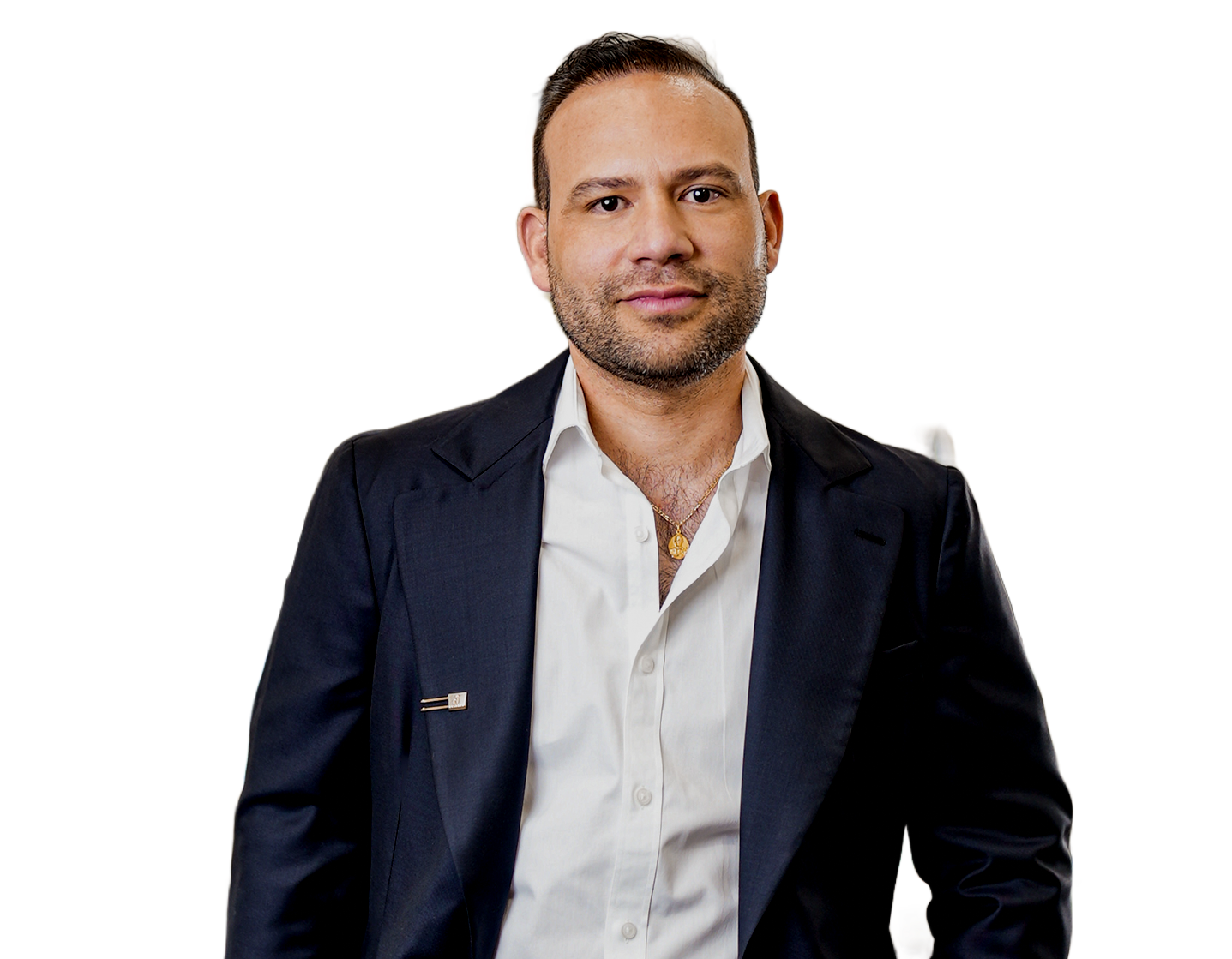 Man in a dark suit and open white shirt, smiling, gold necklace, standing.