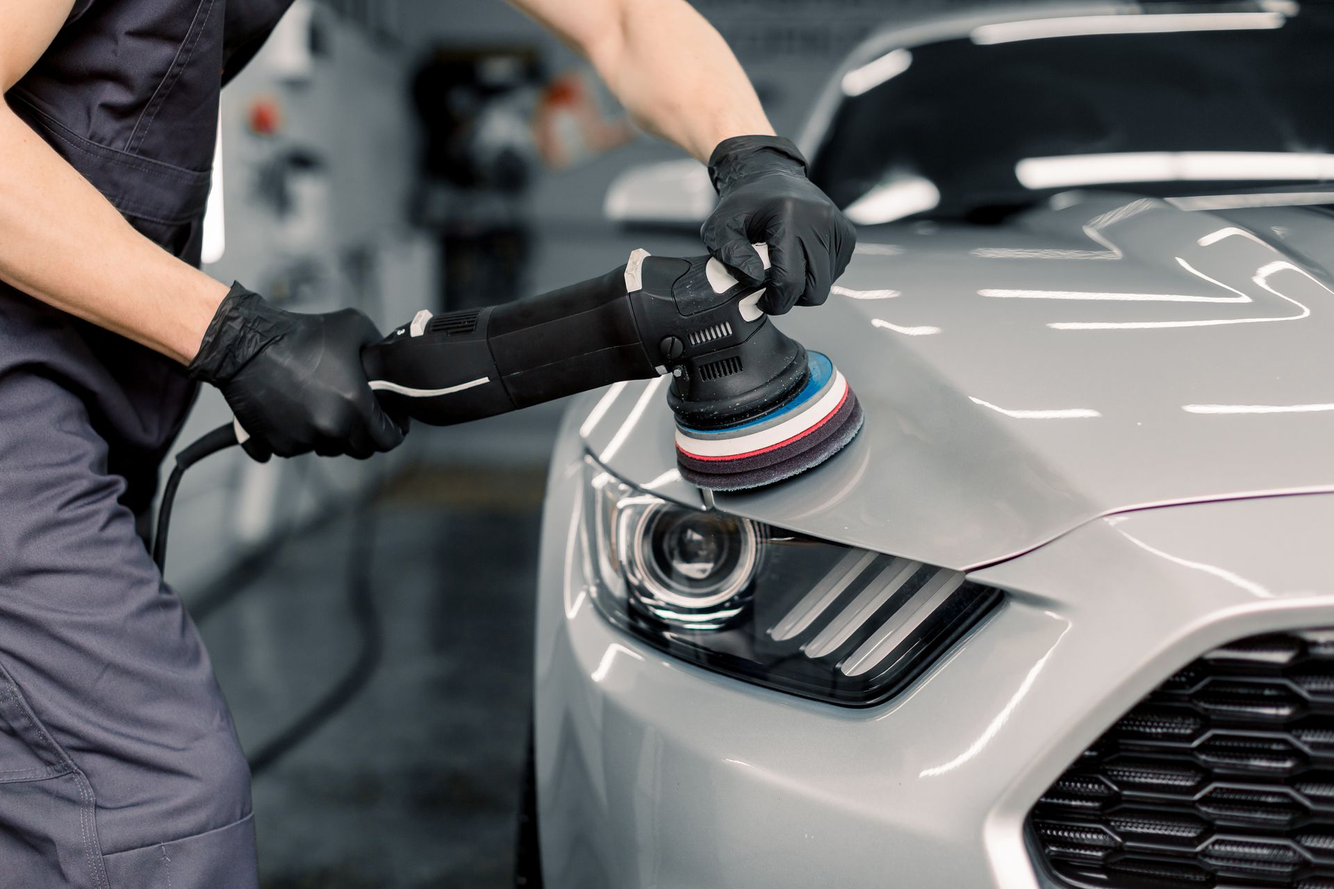 Person using a power buffer on a gray car hood in a garage.