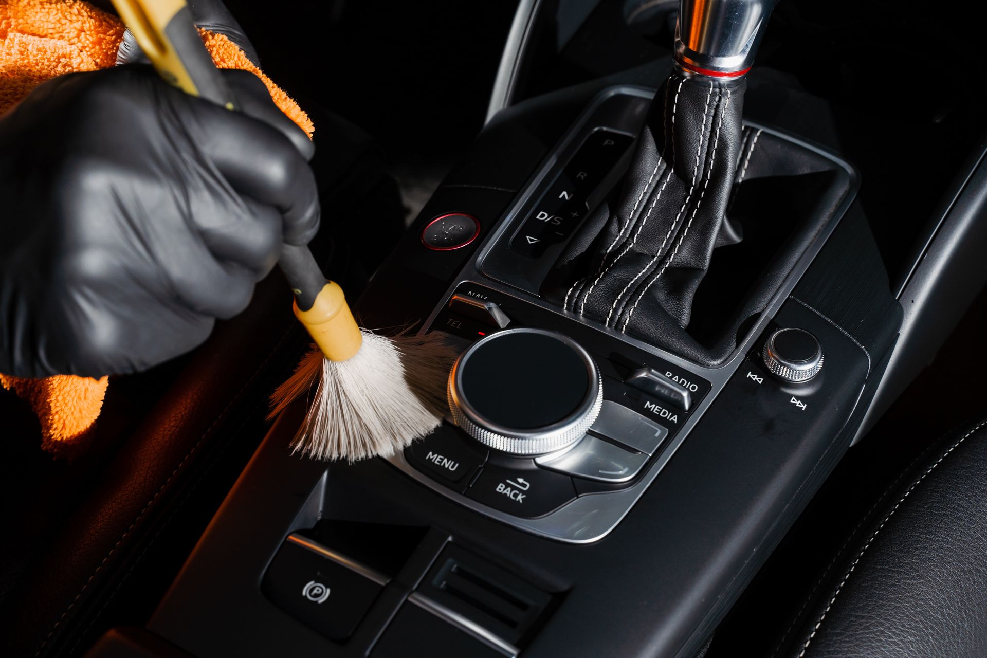 Gloved hand cleaning car's center console with a brush. Orange cloth visible.