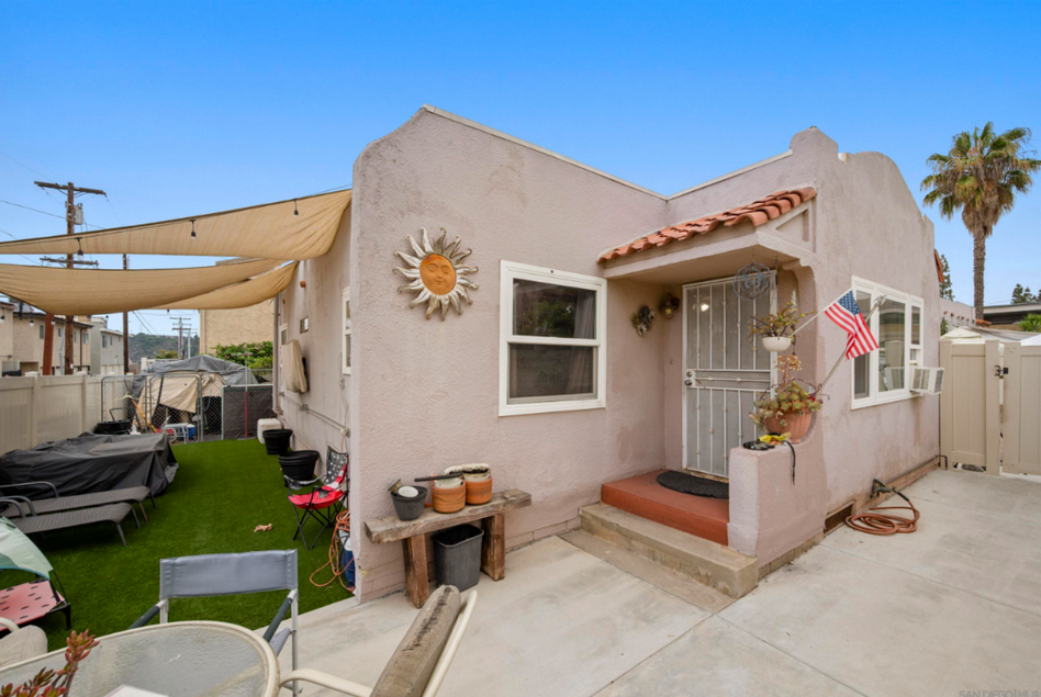 Small beige house exterior with porch, potted plants, and an American flag on a sunny day