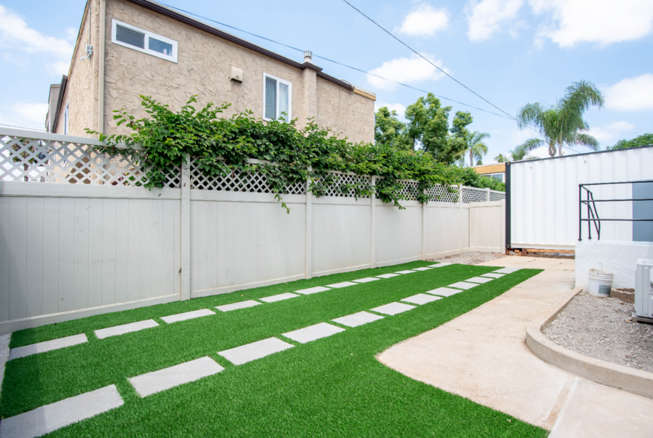 Backyard with green turf path, white fence, stepping stones, and a curved patio beside a beige house