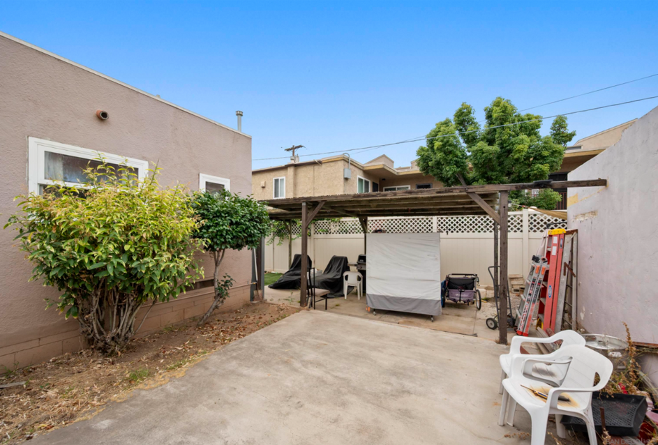 Backyard patio with covered seating, potted shrubs, and white chairs beside beige house walls