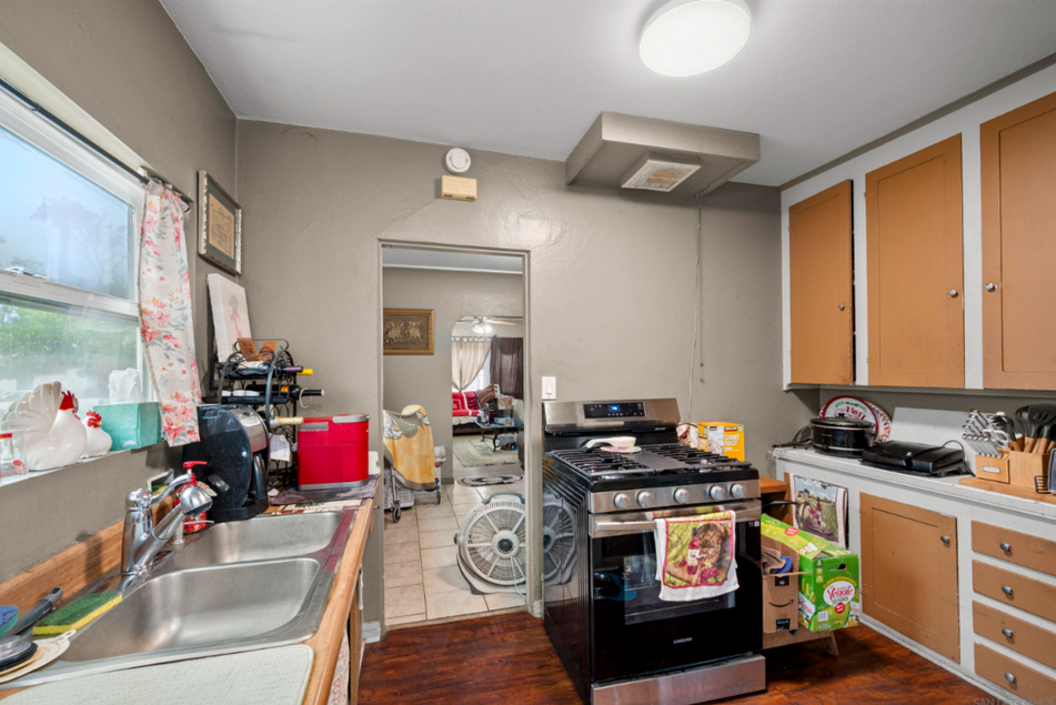 Compact kitchen with sink, stove, orange cabinets, and a doorway to a cluttered back room
