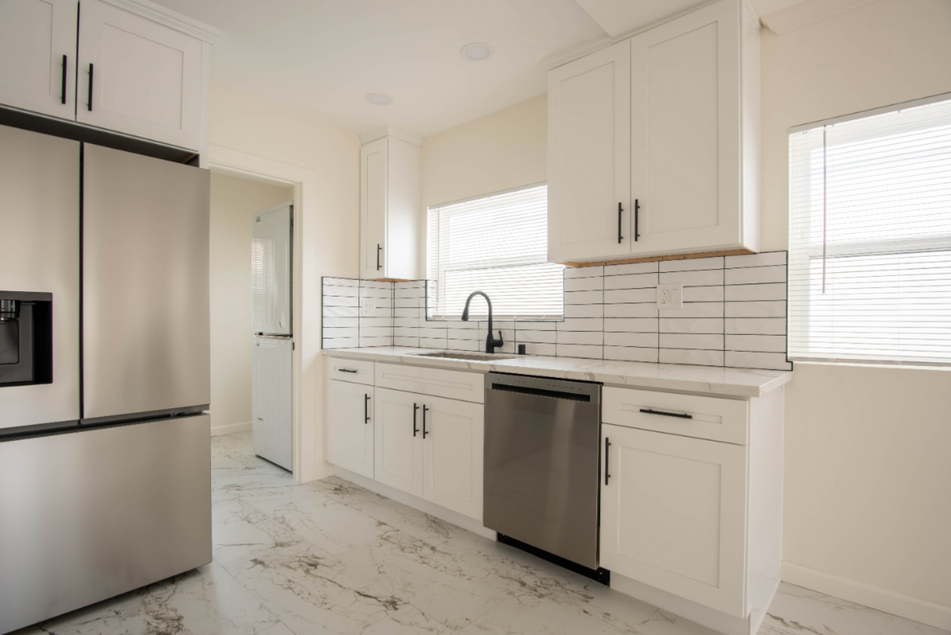 Bright white kitchen with stainless steel appliances, cabinets, and tiled backsplash, lit by windows.