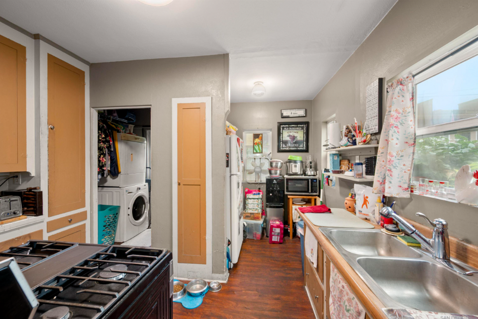 Narrow kitchen with wood cabinets, gas stove, sink, laundry nook, and a cluttered countertop.