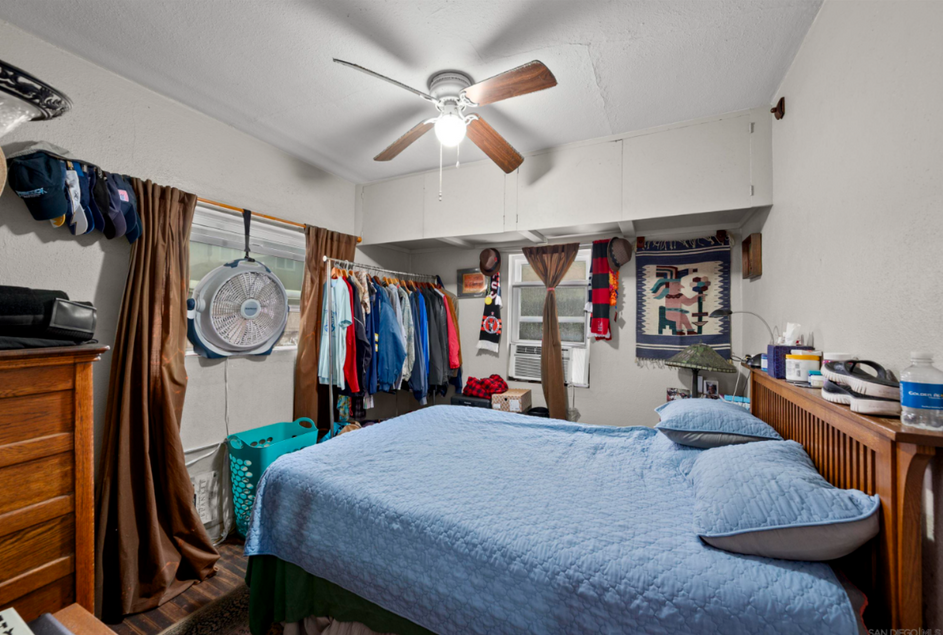 Small bedroom with a blue bedspread, ceiling fan, clothes racks, and a wall clock