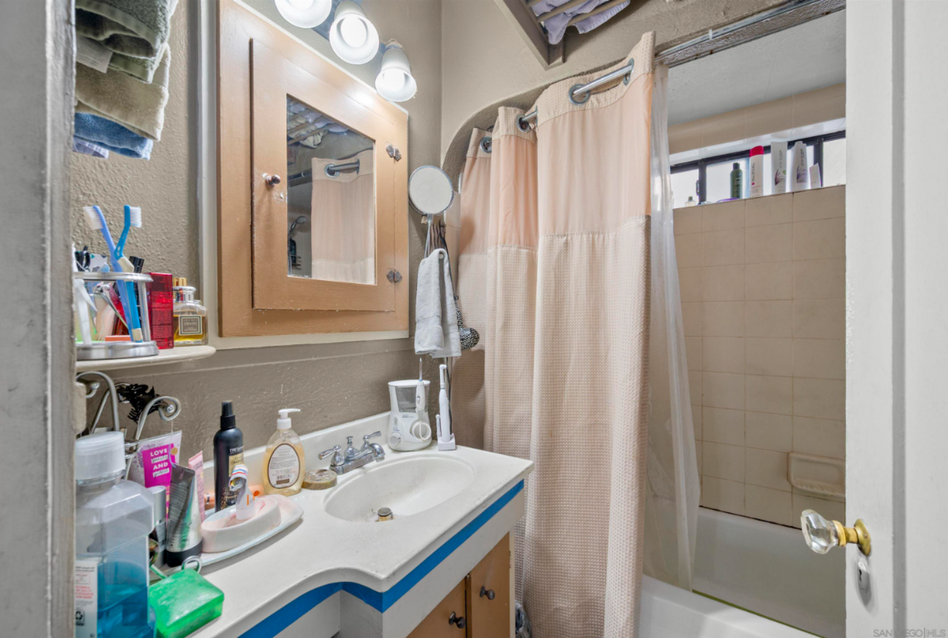 Small bathroom with sink, mirror lights, shower curtain, and tan tiled shower stall.
