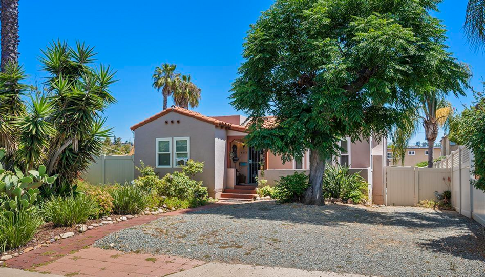 Small stucco house with a red roof, front yard gravel, and a large tree under a clear blue sky.