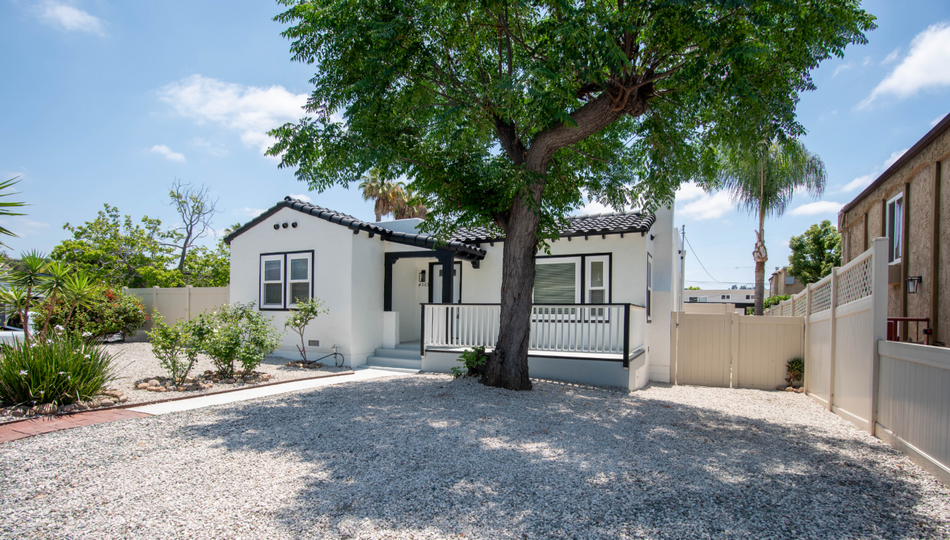 Small white house with a large tree, gravel front yard, and a gated driveway under a clear sky