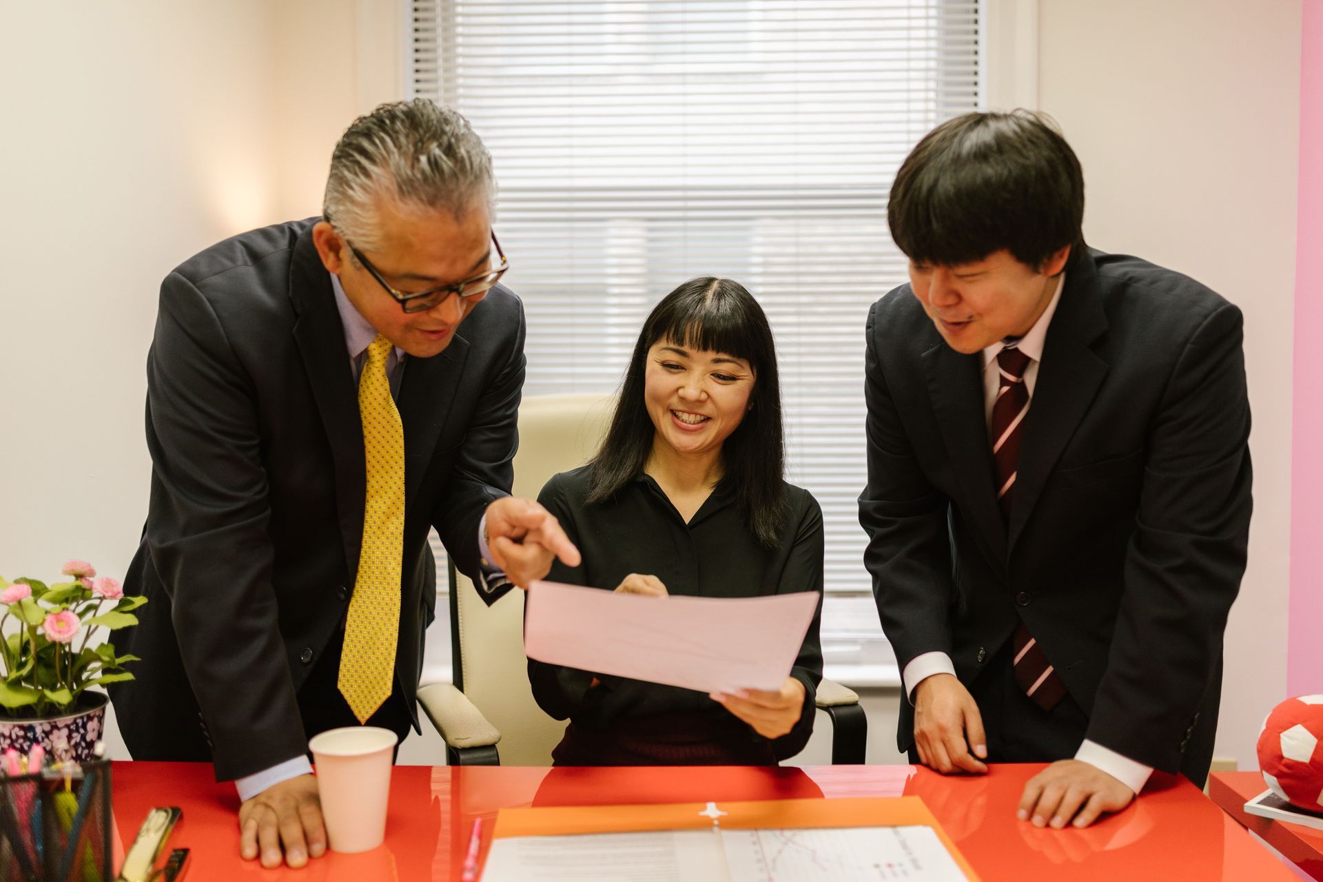 Three people in business attire review a document at a table in a bright office.
