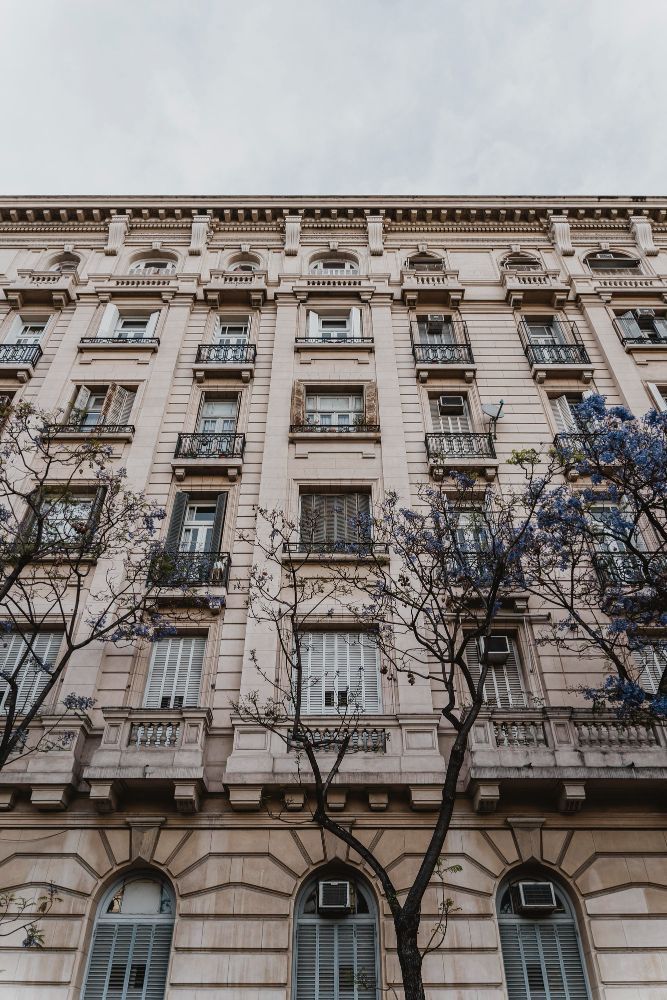 Tall beige apartment building with ornate balconies and bare tree branches in front