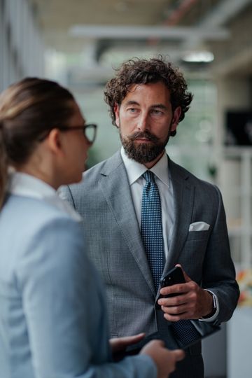 Two professionals talking in an office hallway, one holding a phone and looking attentive.