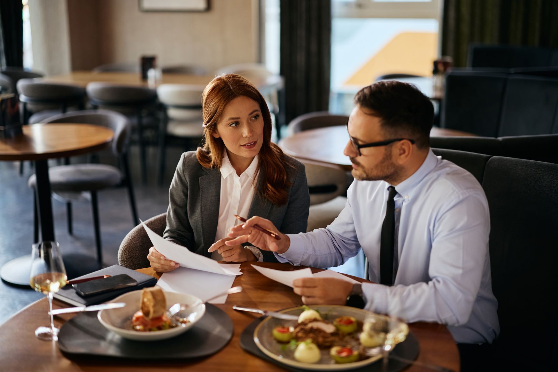 Two coworkers in business attire discussing paperwork at a café table with food and coffee.