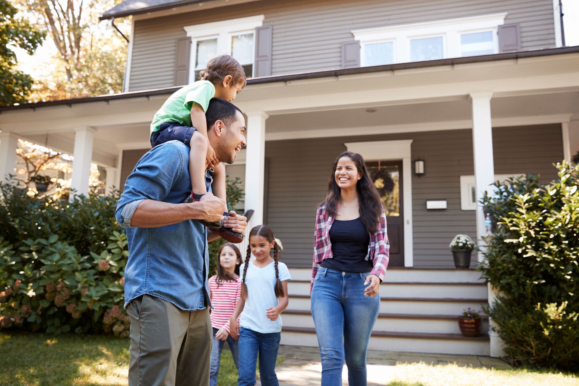 Family standing outside a house, with a child on a man's shoulders and two children nearby, smiling.