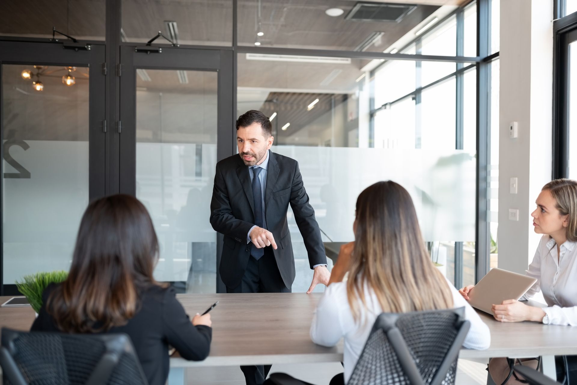 Business meeting in a glass-walled conference room, with a presenter speaking to three seated colleagues.