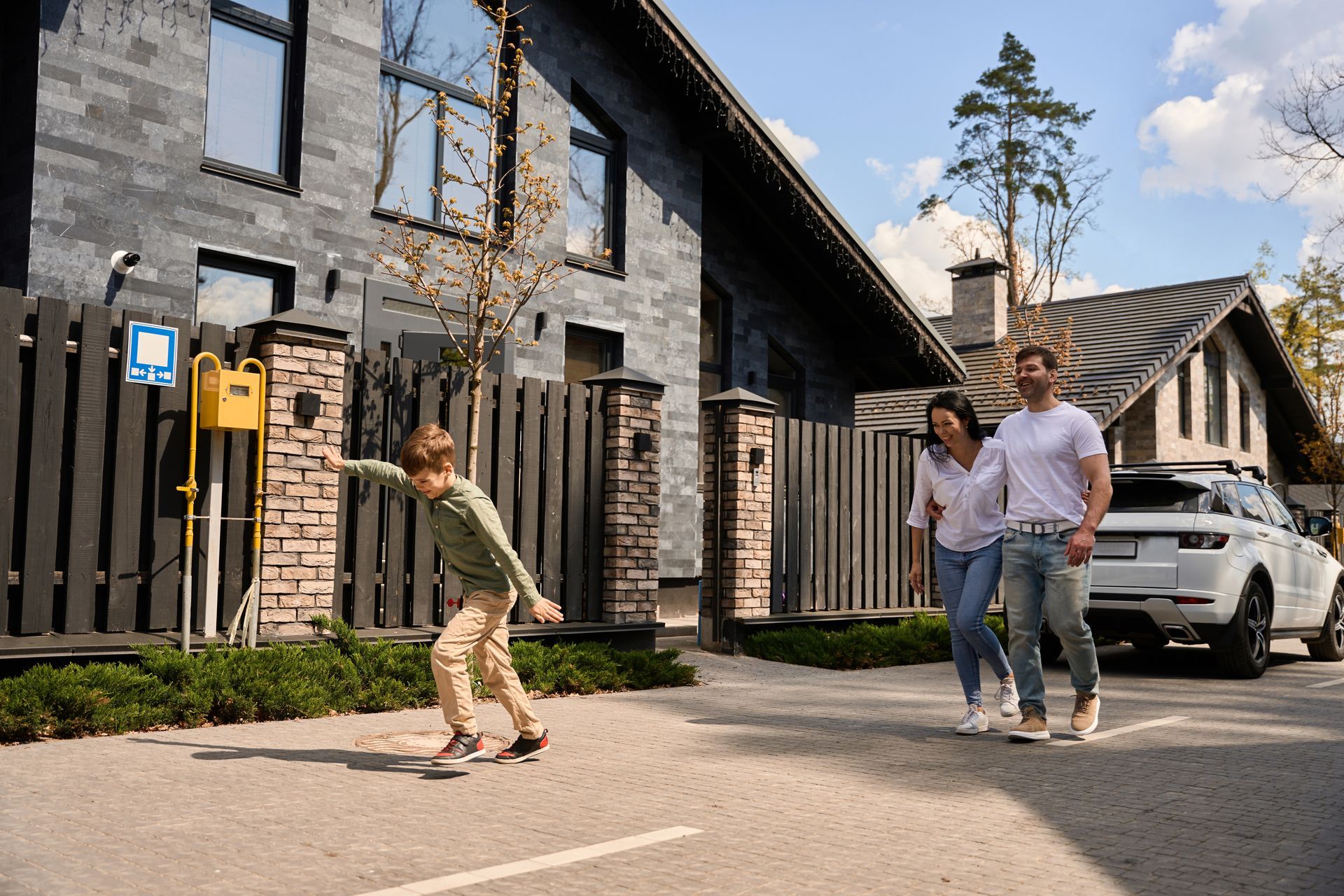 People walking past a stone house; a child runs ahead near a parked white SUV.