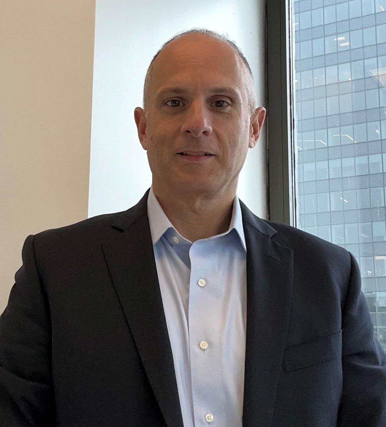 Man in a dark blazer and light blue shirt standing indoors near a window with city buildings behind him
