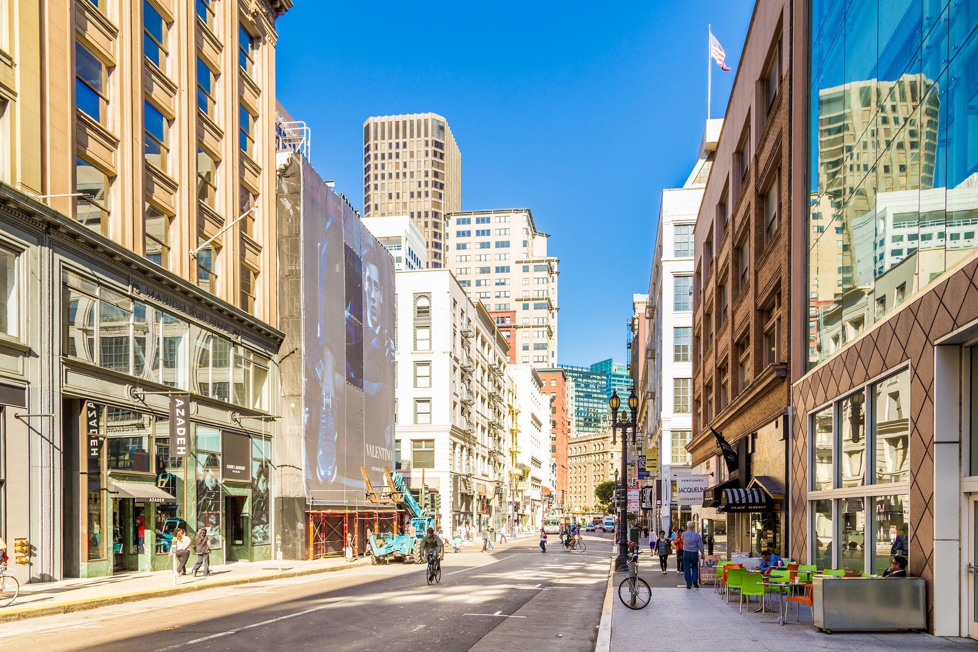 Sunny city street lined with tall buildings and storefronts, with a few pedestrians and cyclists.