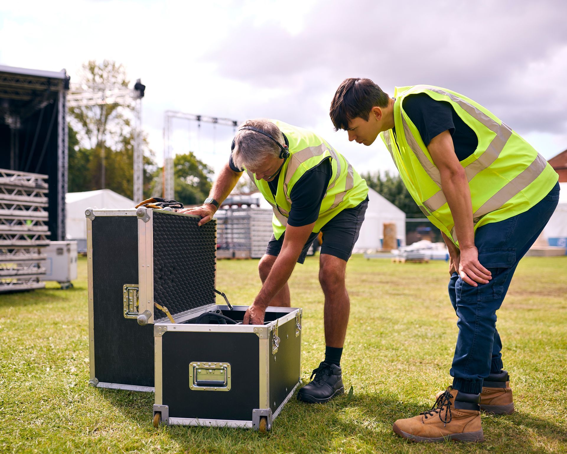 Two workers in neon safety vests unload equipment from black road cases on a grassy field.