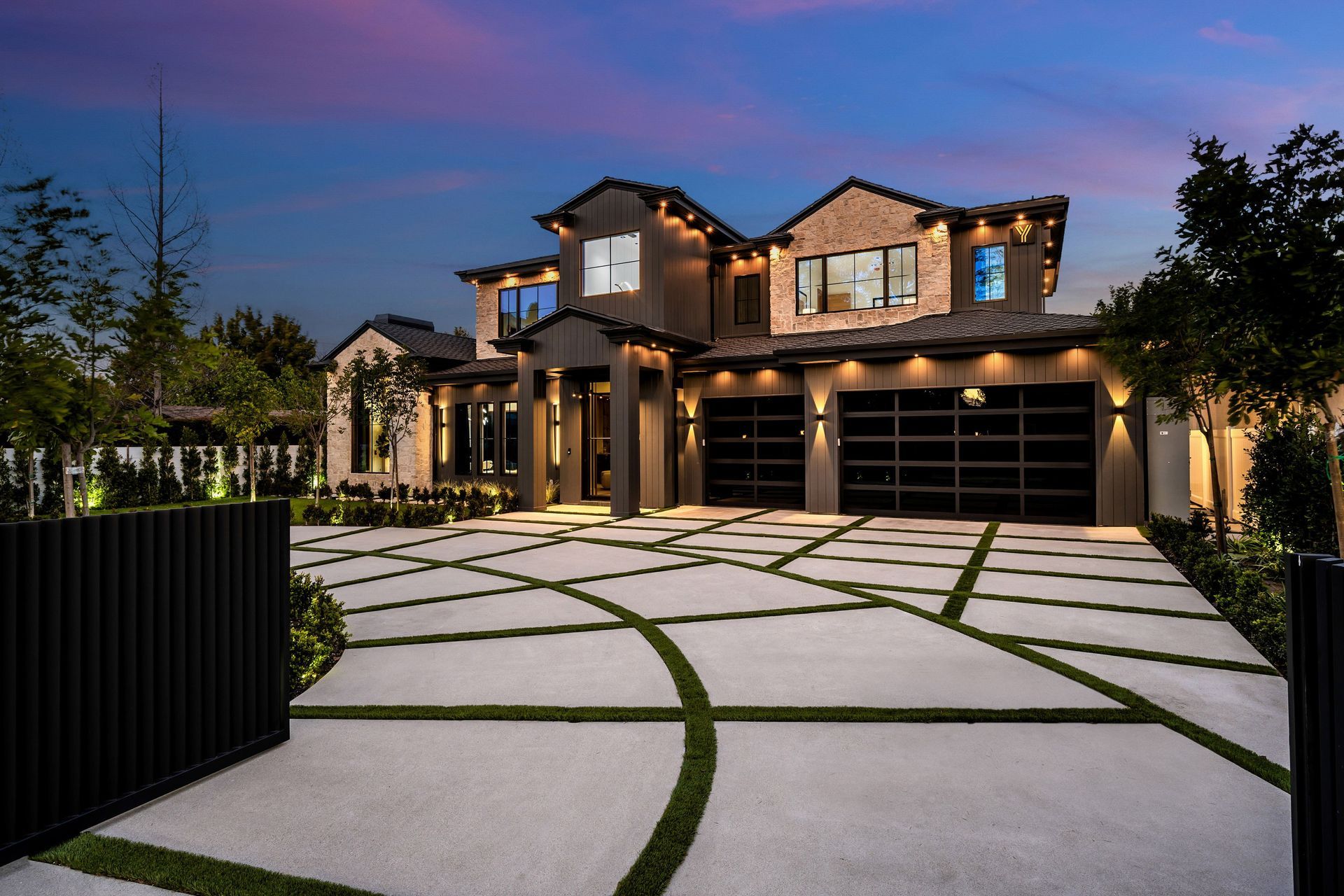 Modern two-story house at dusk with lit windows and a patterned driveway behind a black gate