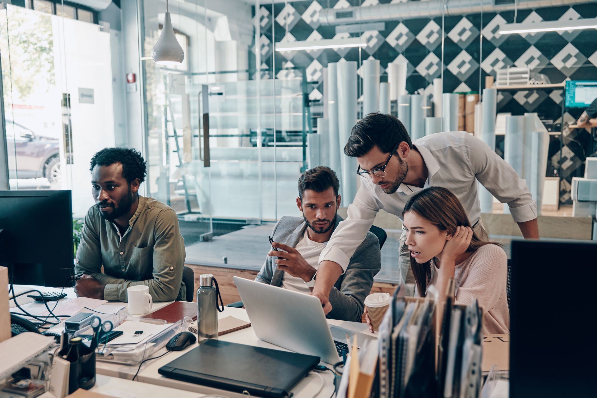 Four coworkers collaborating around a laptop in a modern glass office