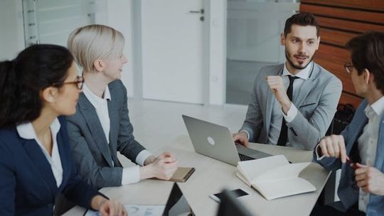 Four coworkers in a meeting around a laptop, discussing documents in a bright office.