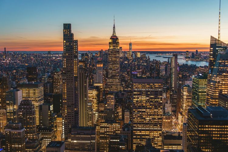 City skyline at sunset with lit skyscrapers and a glowing orange horizon over the water