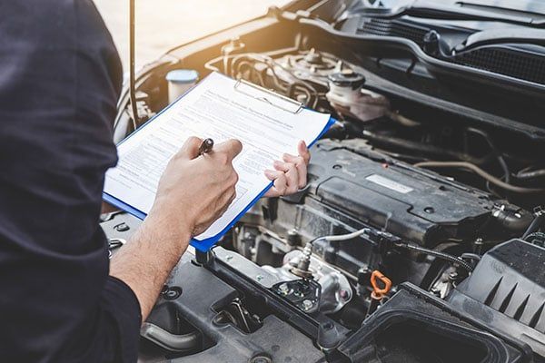 A man is writing on a clipboard while looking under the hood of a car. | Lex Brodies 