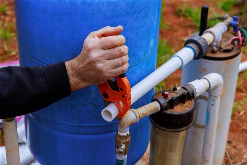 Person cutting white PVC pipe with orange cutter near blue and beige water tanks.