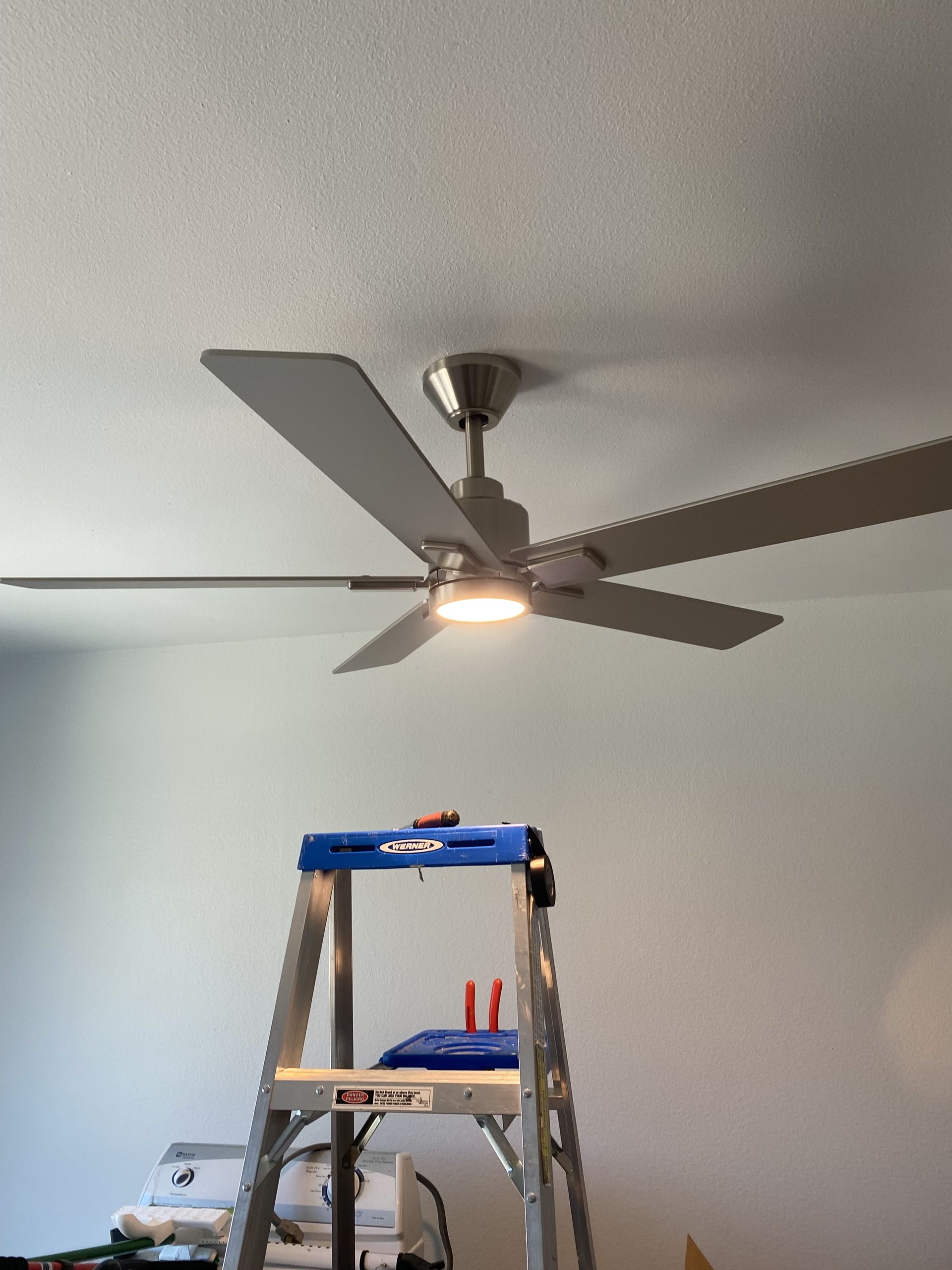 Ceiling fan above a step ladder in a room with white walls and a lit bulb.