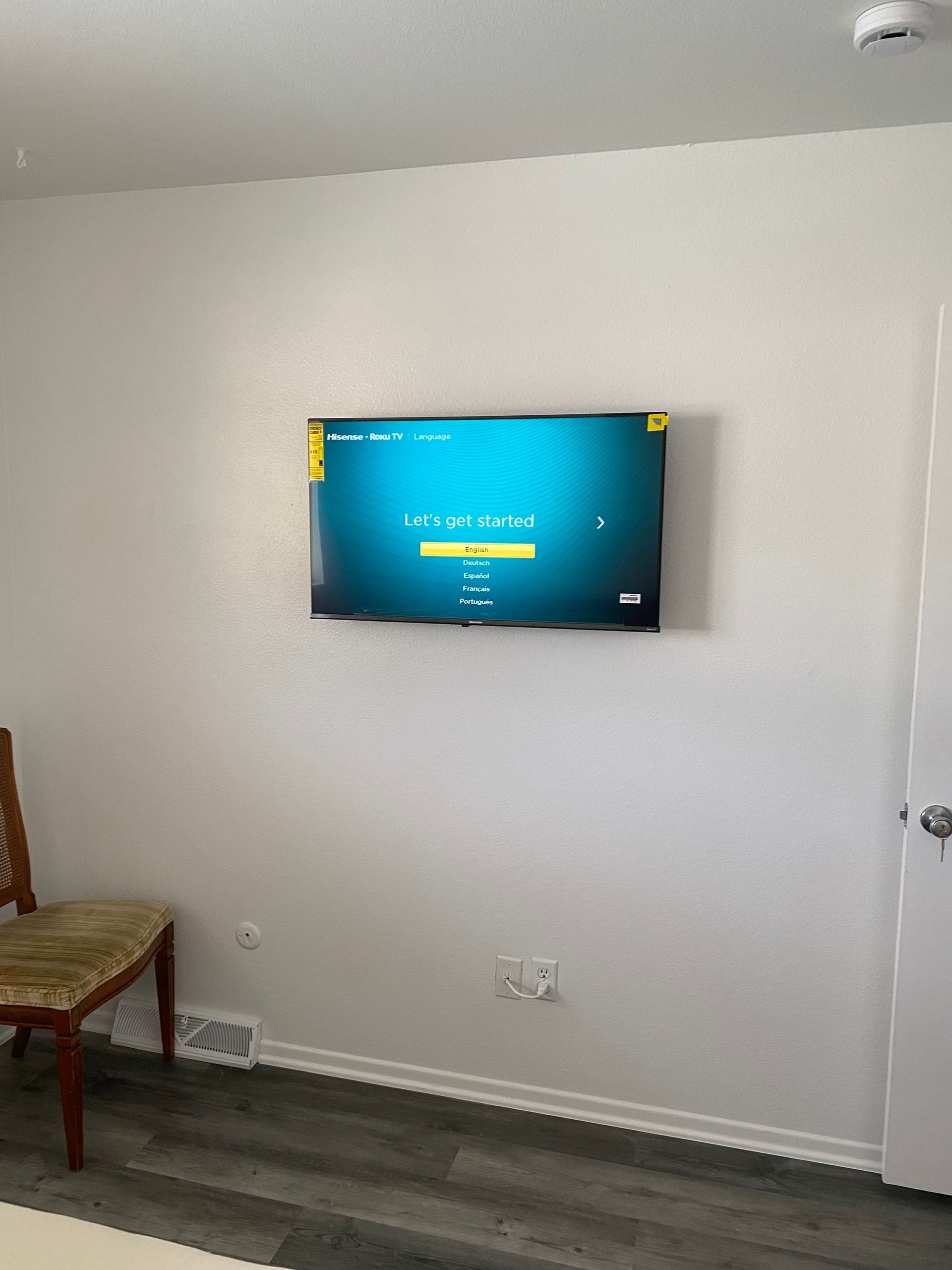Small room with a wall-mounted TV displaying a blue screen, a chair on the left, and a white wall.
