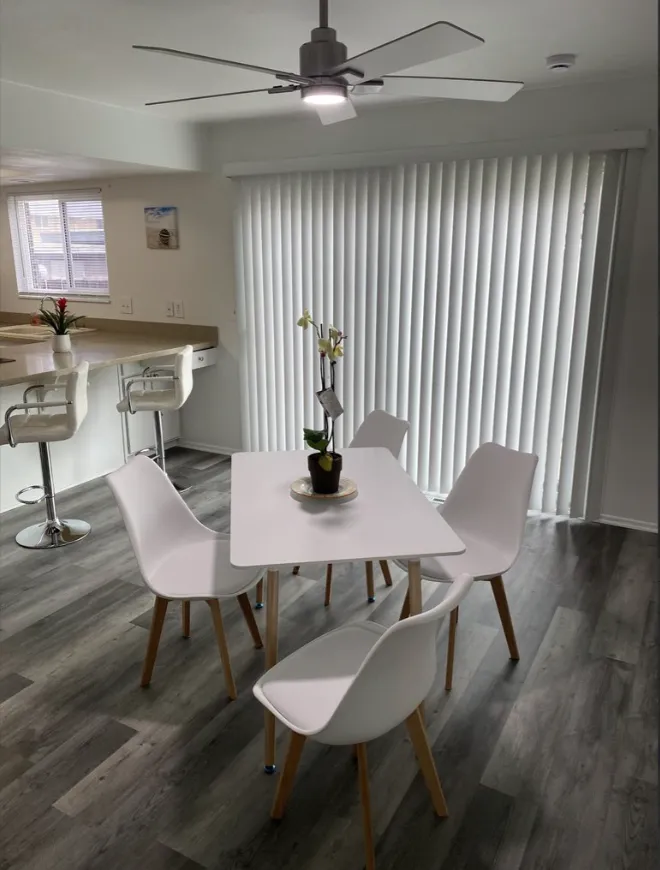 Modern dining area with white chairs, square table, ceiling fan, and large vertical blinds by a kitchen bar.