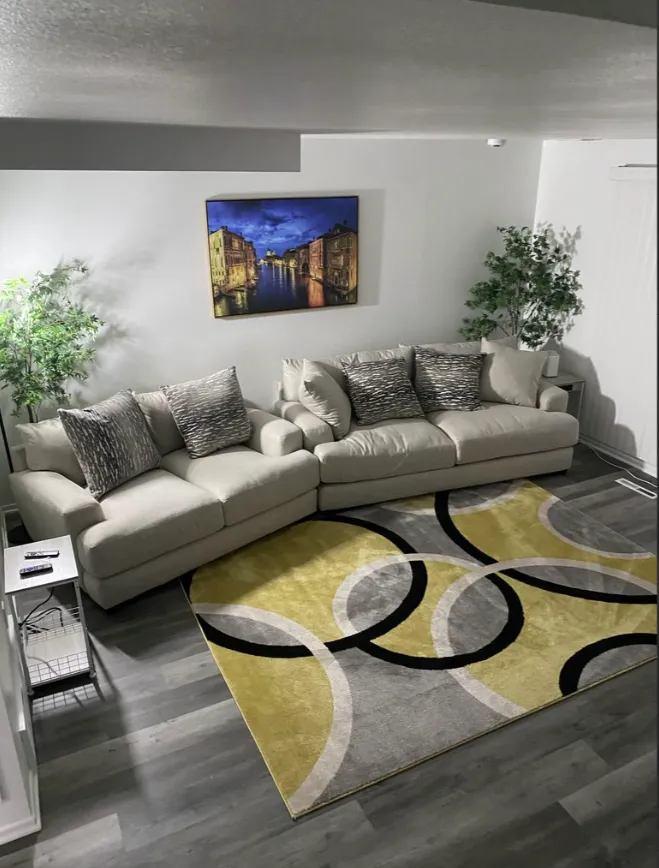 Modern living room with beige sectional sofa, patterned rug, wall art, and potted plants.