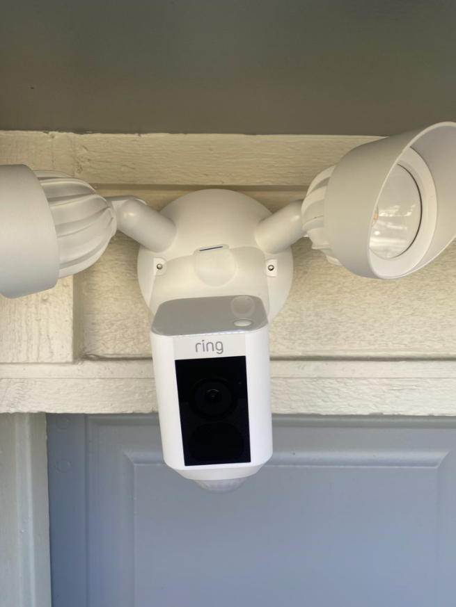 Ring security camera mounted above a doorway with two floodlights