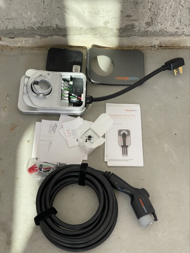 EV charger kit with coiled black cable, gray charging unit, plug, and instruction booklet on a concrete floor