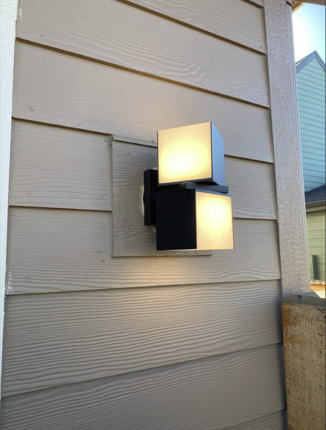 Two-tone outdoor wall sconce on beige vinyl siding, lit against a house exterior.