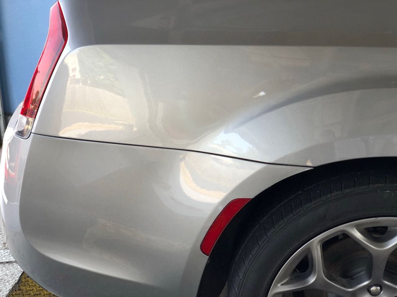 Close-up of a silver car’s rear quarter panel and wheel with a red reflector, showing a dent and scratch.