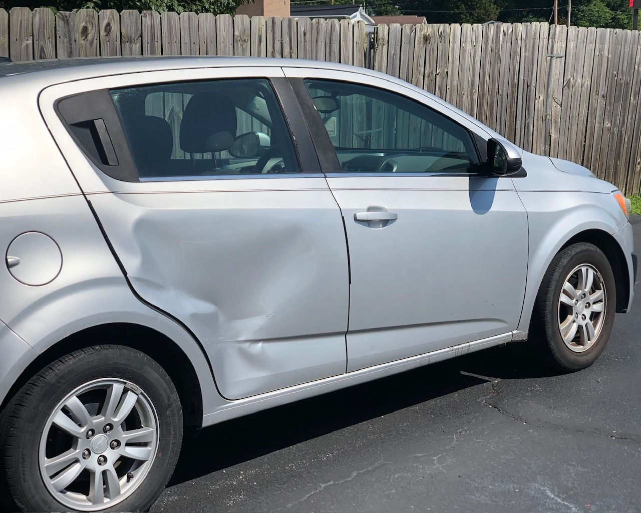 Silver hatchback car with a large side dent, parked on a driveway by a wooden fence.