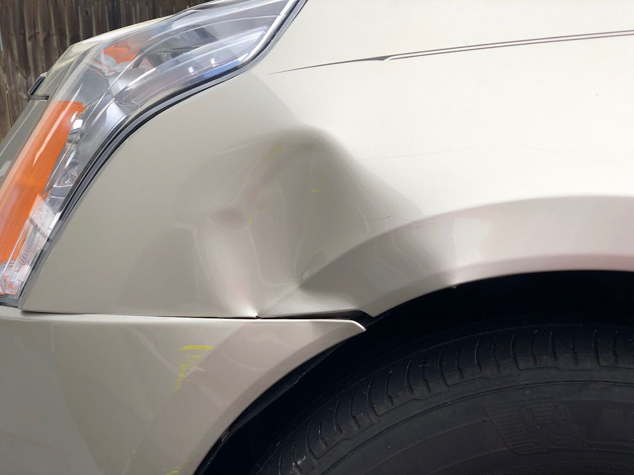 Close-up of a beige car’s front fender with a dent near the wheel well and headlight.