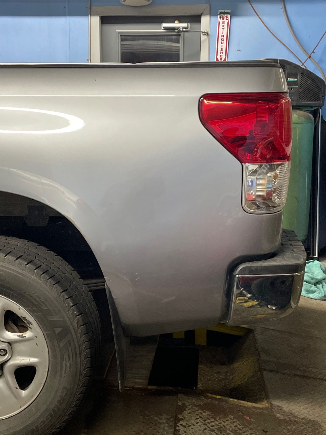 Rear side of a silver pickup truck parked near a building, showing the taillight and bumper.