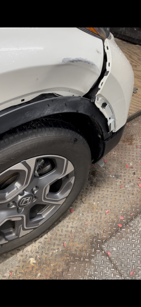 Close-up of a white car’s front wheel and fender with visible body damage and a gap at the panel seam.