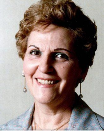 Woman smiling, wearing pearl earrings, light-colored patterned blazer; short, curly hair.